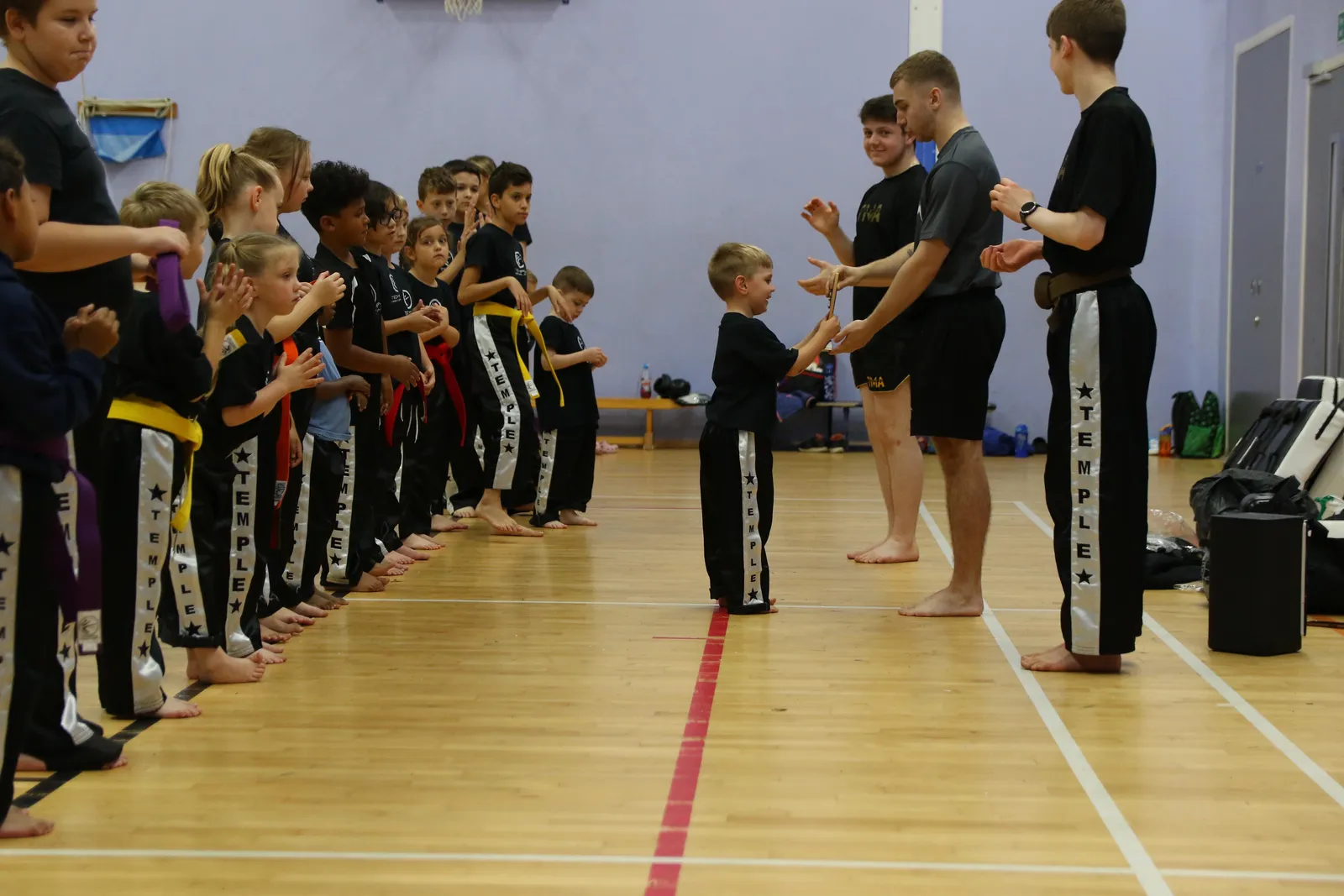 A line of kids in belts facing their coach in a sports hall