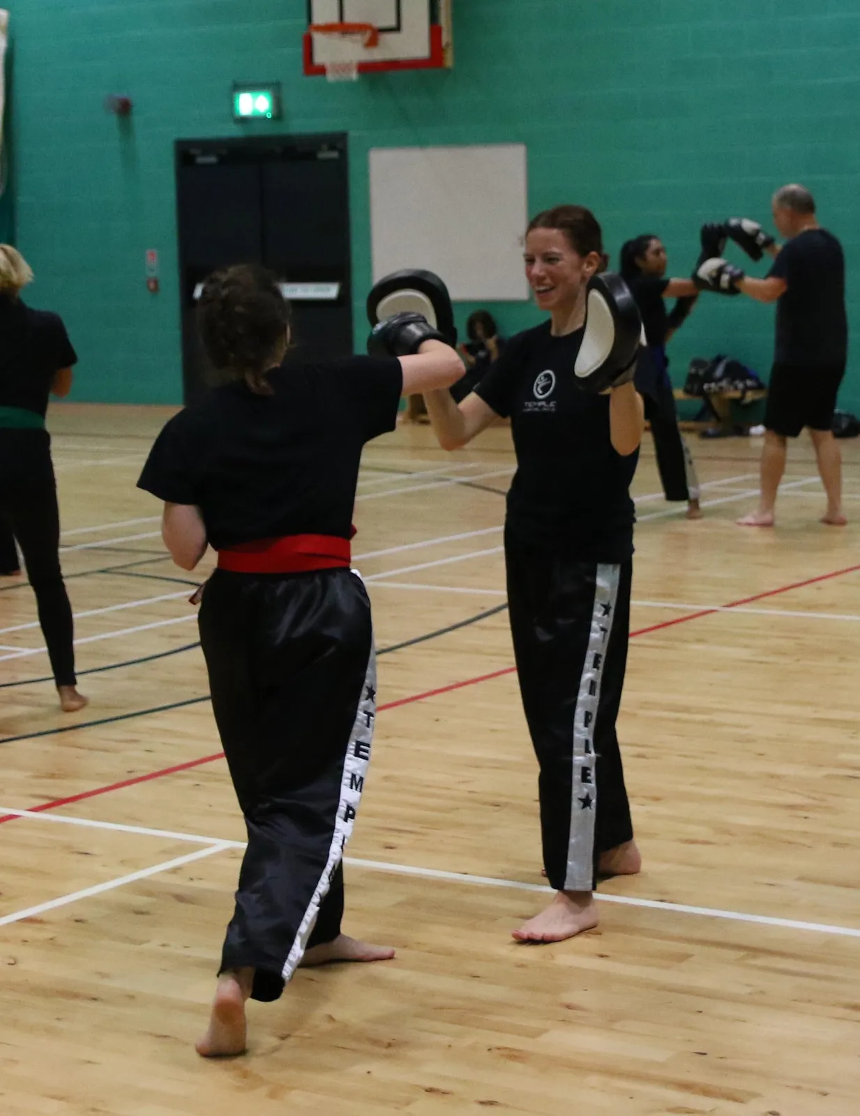 A smiling woman holding focus pads as her partner throws a punch, with other adults training behind them